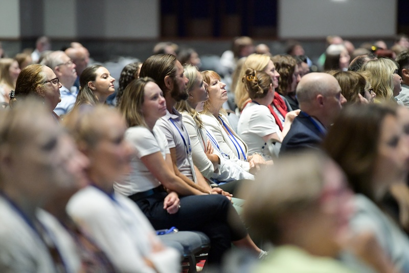 image shows an audience looking toward a stage in a convention hall